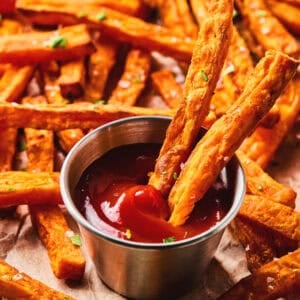 Close up of two sweet potato fries in a container of ketchup with more sweet potato fries in the background.