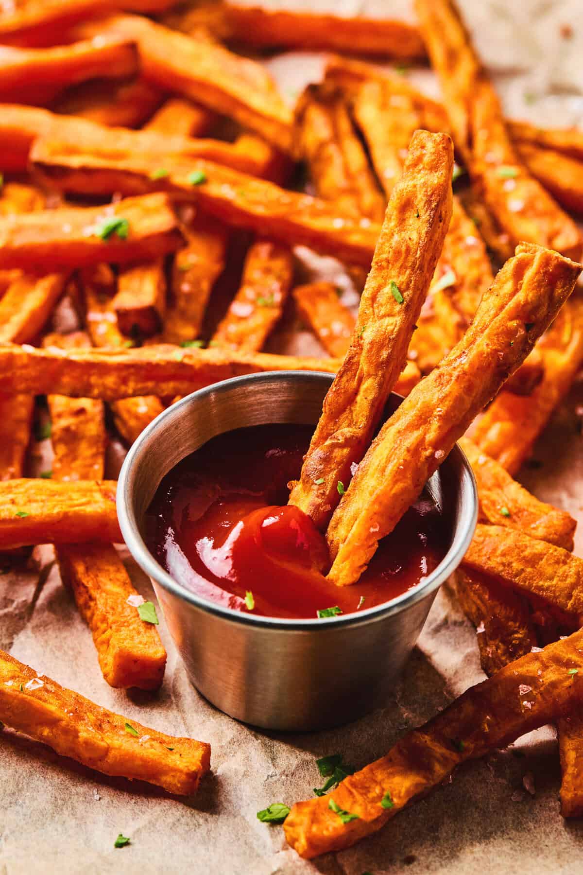 Close up of two sweet potato fries in a container of ketchup with more sweet potato fries in the background.