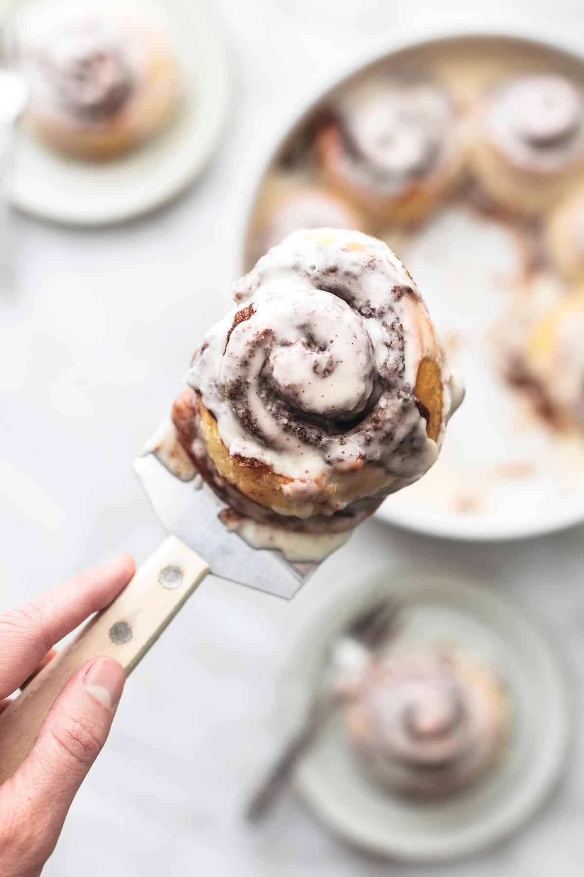 top view of a frosted cinnamon roll on a spatula above a round dish and plates of more rolls.