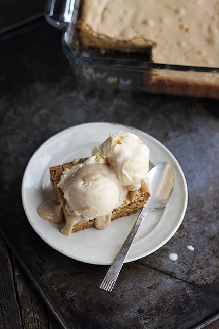 blondie sundaes with hot maple cream sauce on white plate with a spoon on a baking sheet with the blondie brownies in a baking pan in the background.