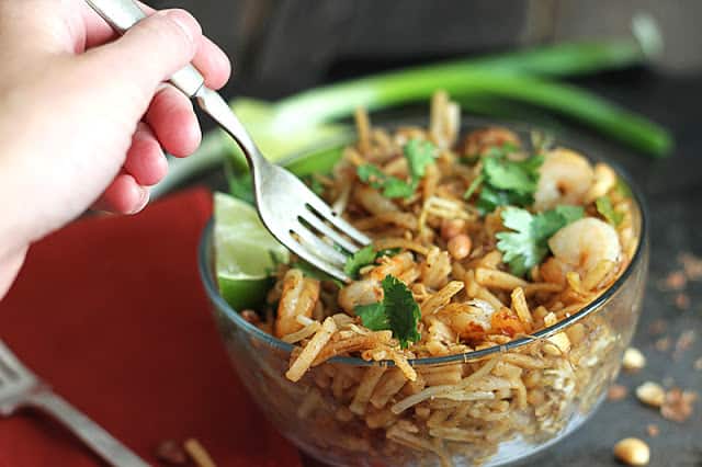 a hand taking some shrimp pad Thai with a fork out of a glass bowl.