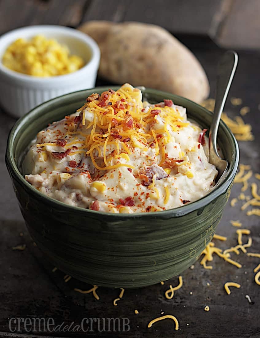 chowder in a green bowl with a spoon on a baking sheet with corn in a bowl and a potato in the background.