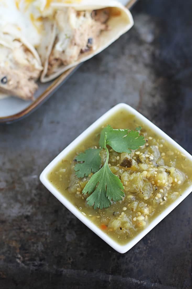 salsa verde in small white square bowl with enchiladas on a plate faded in the background.