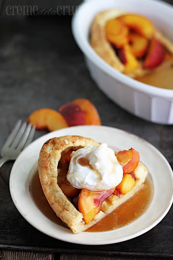 puff pancake on a plate topped with syrup and whipped cream on a baking sheet with a fork, slices of peaches and the rest of the puff pancake in serving bowl.