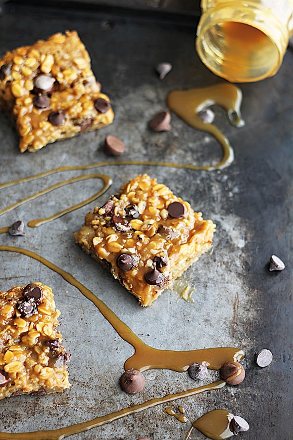 caramel chocolate chip oat bars on a baking sheet with tipped mason jar and caramel drizzled over the sheet with chocolate chips.