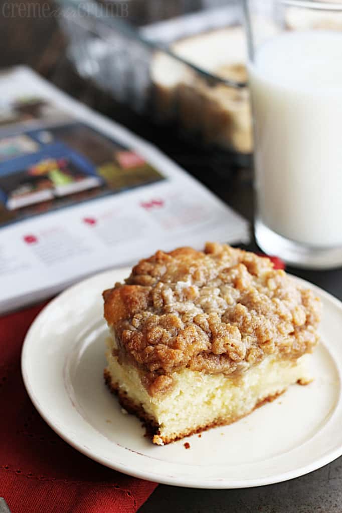 a piece of crumb cake on a plate with a glass of milk and a magazine faded in the background.
