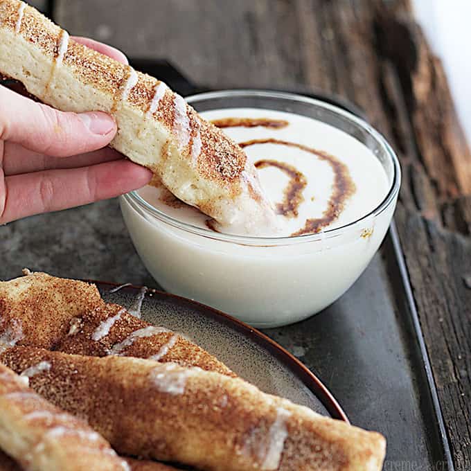 a hand dipping a cinnamon roll dipper in glaze sauce in a bowl with cinnamon roll dippers on a plate on the side.
