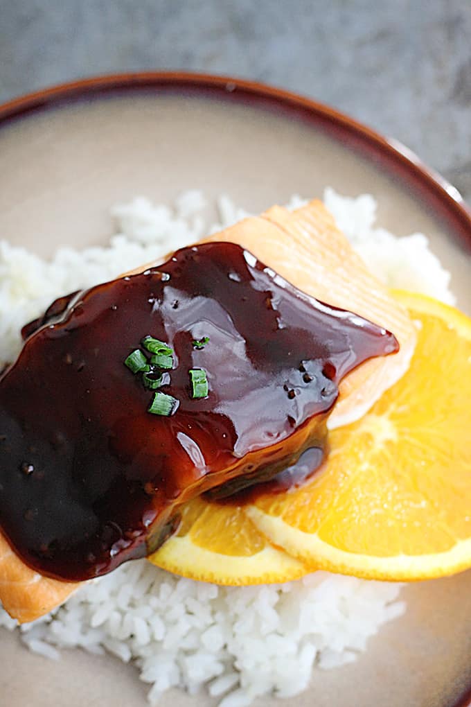 top view of orange teriyaki salmon on orange slices on rice on a plate.