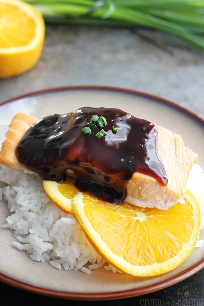 orange teriyaki salmon on orange slices on rice on a plate with a half an orange and a bundle of green onions faded in the background.