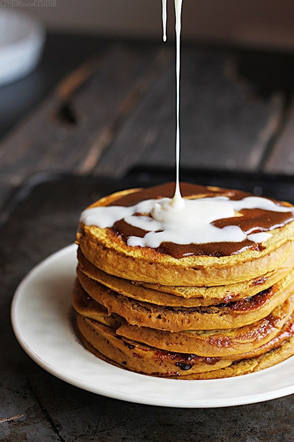 pumpkin cinnamon roll pancakes on a plate with cream cheese vanilla glaze being drizzled on top.