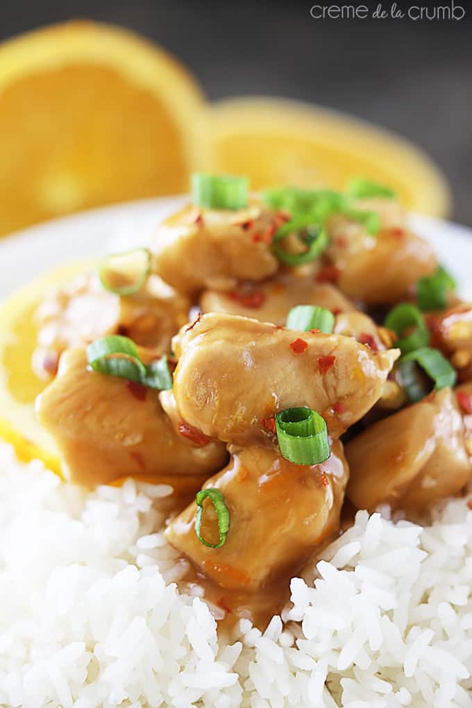 close up baked orange chicken on rice in a bowl with two orange halves faded in the background.