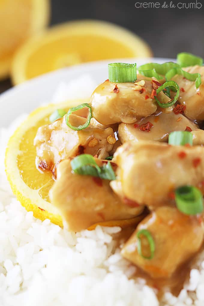 close up side view of baked orange chicken on rice in a bowl with two halves of an orange faded in the background.