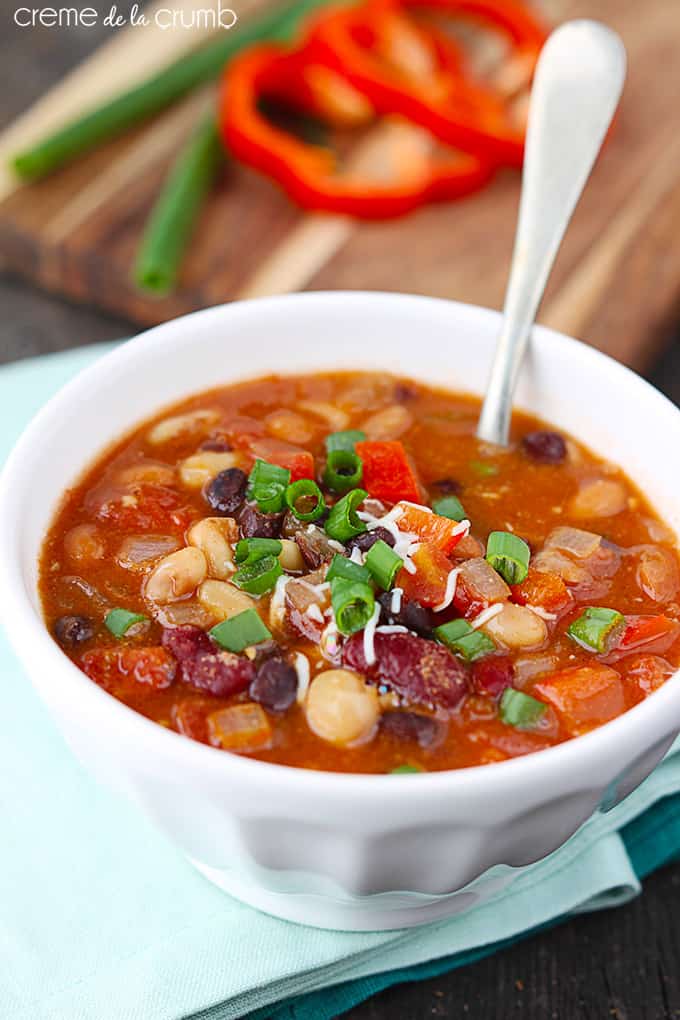 five bean vegetarian Chili with a spoon in a bowl on a green cloth napkin with slices of red pepper and green onions on a wooden cutting board faded in the background.