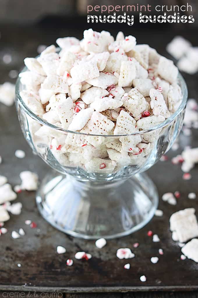 peppermint crunch muddy buddies in a glass pedestal bowl on a baking sheet with more peppermint pieces and muddy buddies and the title of the recipe written on the top right corner of the image.