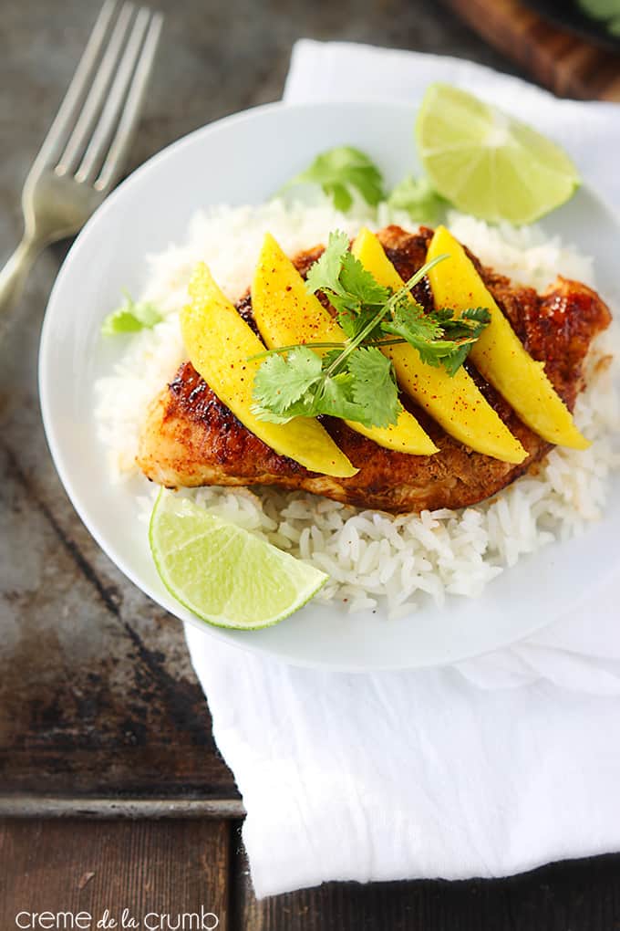 sweet paprika chicken on rice topped with mango slices and cilantro leaves with slices of a lime on the side of the plate on a white cloth napkin and a fork on the side.