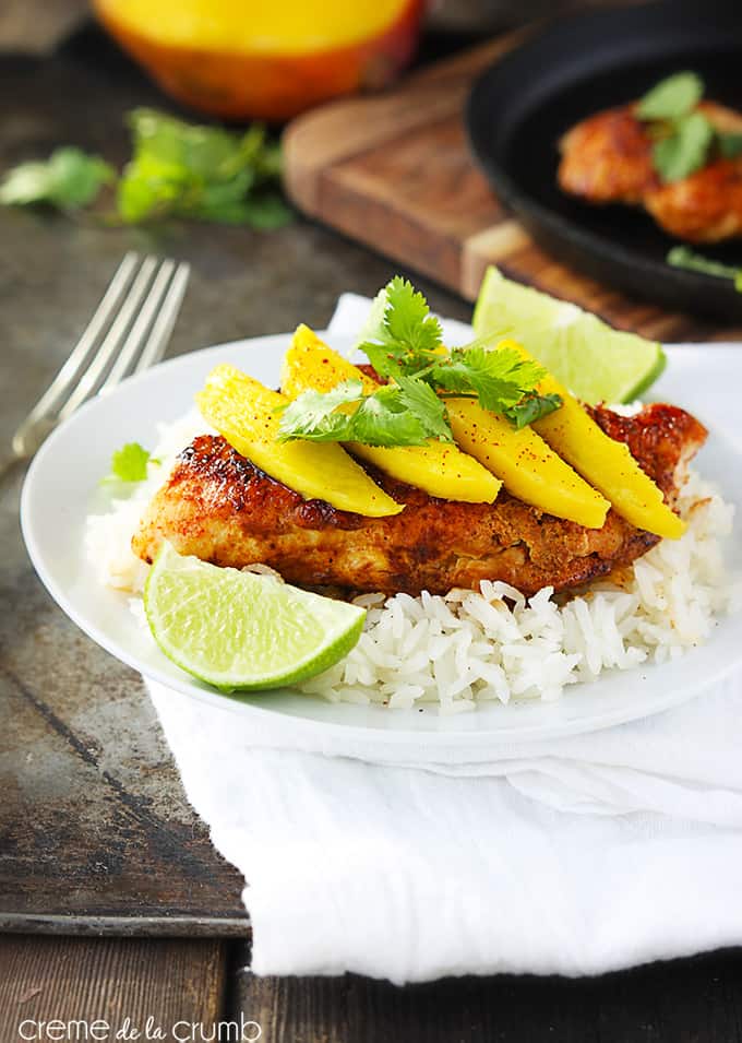 sweet paprika chicken on rice topped with mango slices and cilantro leaves with slices of a lime on the side of the plate on a white cloth napkin with a fork on the side with a cutting board and ingredients faded in the background.