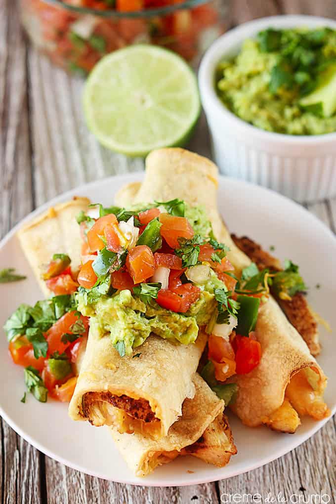 baked cheesy chicken taquitos on a plate with guacamole and pico de Gallo on top with the toppings and a half a lime in bowls faded in the background.