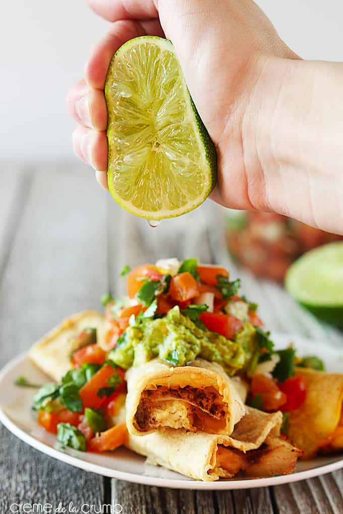 baked cheesy chicken taquitos topped with guacamole and pico de Gallo on a plate with a hand squeezing half of a lime on top.