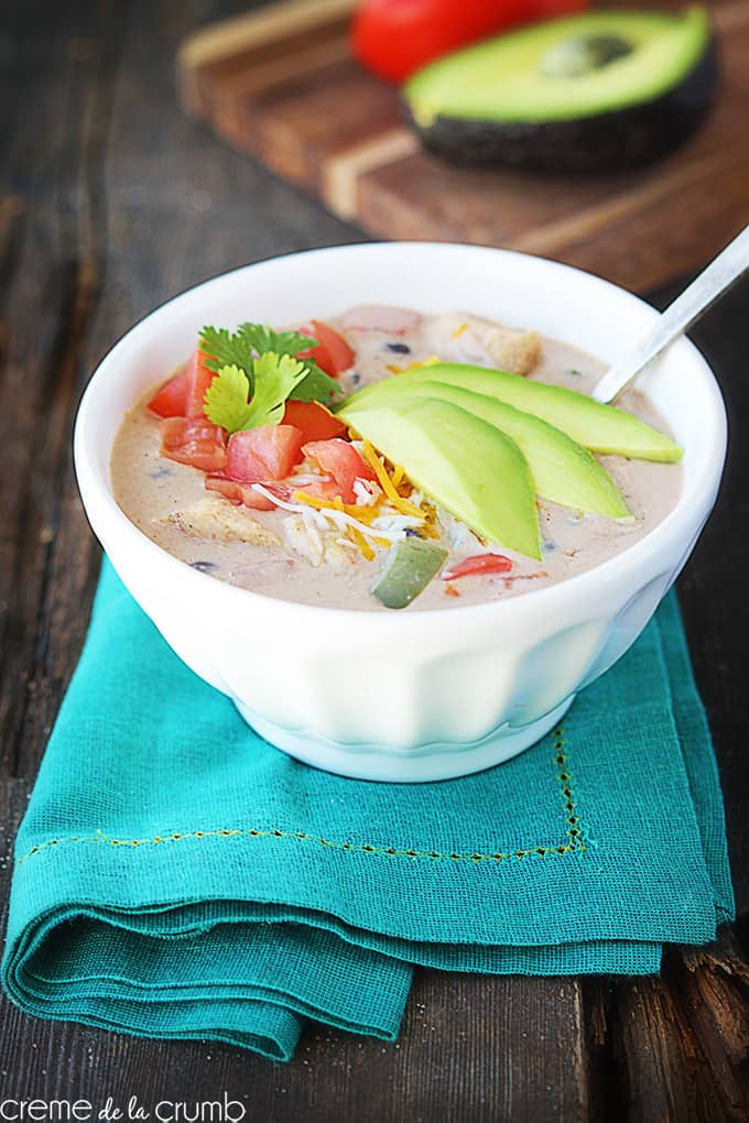 chicken fajita soup with a spoon in a bowl on a blue cloth napkin with a wooden cutting board with half of a tomato and avocado on it faded in the background.