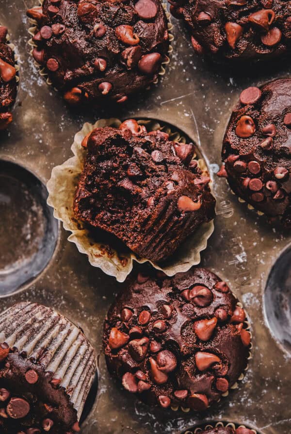 Overhead of chocolate muffins in a muffin tin with a bite out of one muffin.