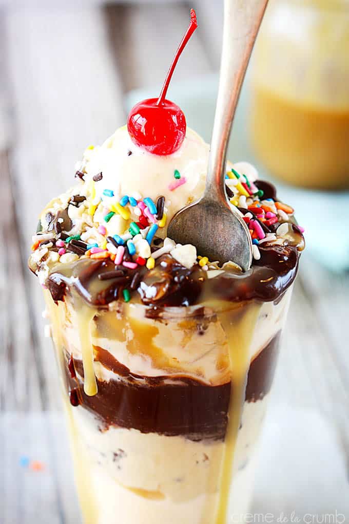 close up of chocolate chip cookie dough hot fudge sundae topped with a cherry in a tall glass with a spoon.