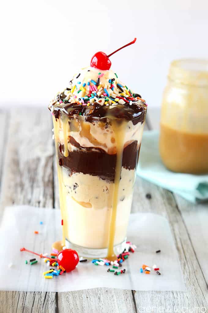 chocolate chip cookie dough hot fudge sundae topped with a cherry in a tall glass with caramel dripping down the side.