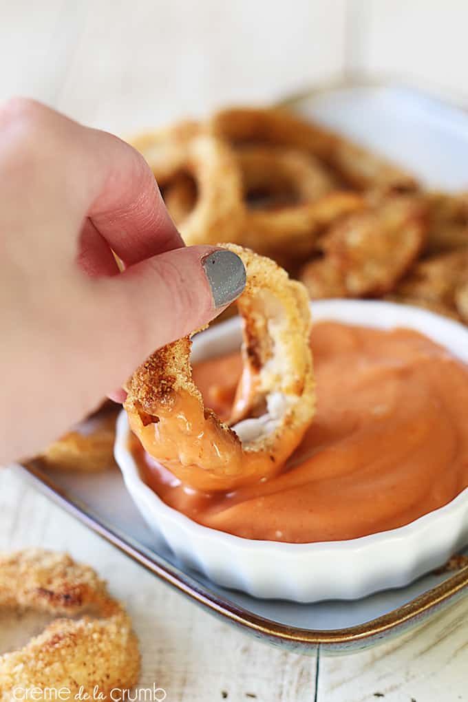 a hand with one of the oven baked onion rings from a tray dipping it in sauce.