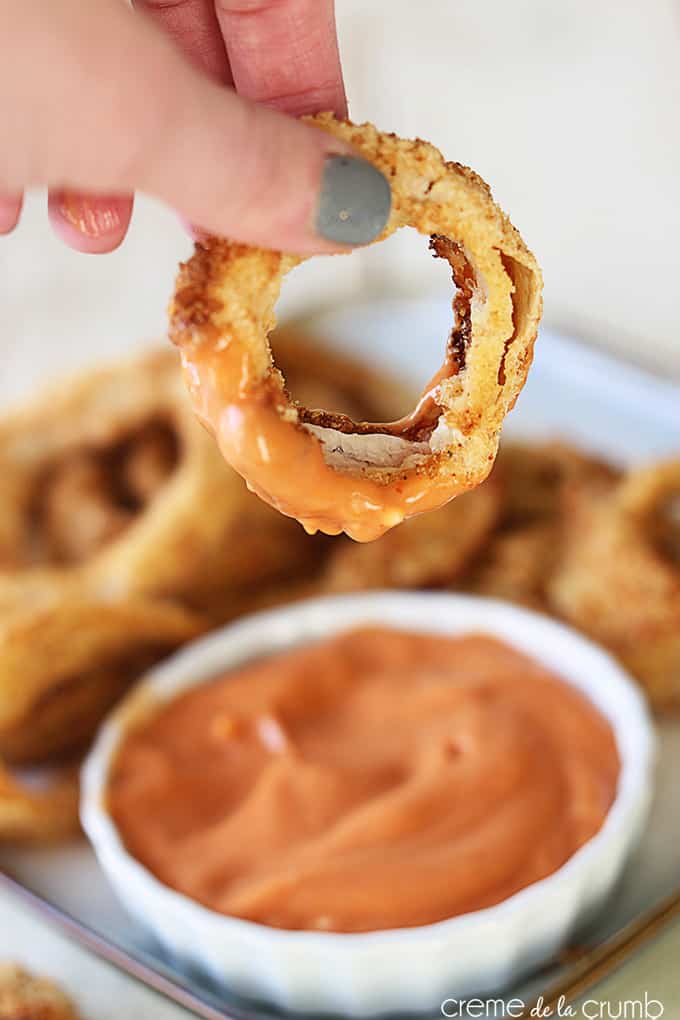 a hand holding a oven baked onion ring dipped in sauce with the sauce and onion rings on a tray faded in the background.