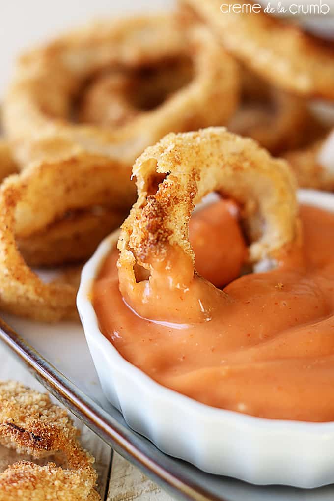 close up of oven baked onion rings on a tray with sauce in a bowl and an onion ring dipped in the sauce bowl.