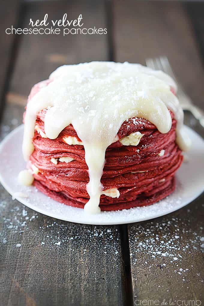 red velvet cheesecake pancakes on a plate topped with cream cheese icing with the title of the recipe written on the top left corner of the image.