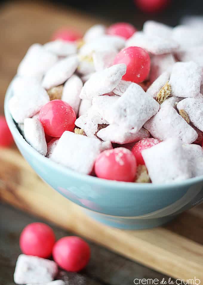 close up of strawberry cheesecake muddy buddies in a bowl on a cutting board with pink candy balls and muddy buddies.