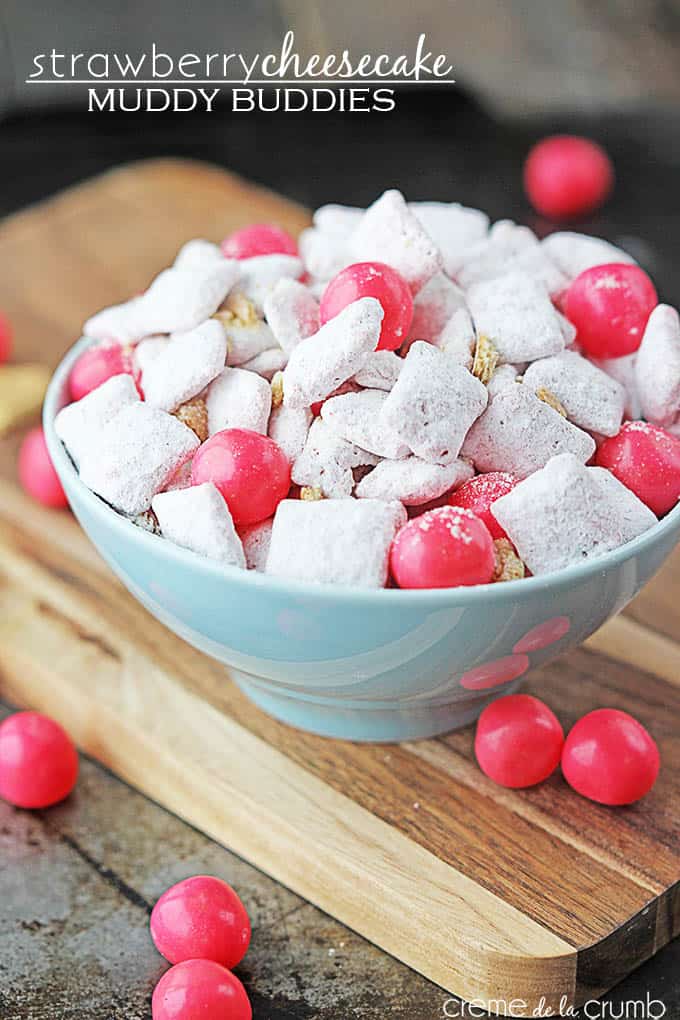 strawberry cheesecake muddy buddies in a bowl on a cutting board with pink candy balls with the title of the recipe written on the top left corner of the image.