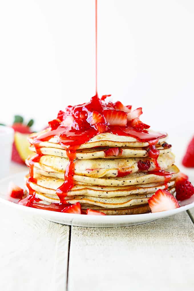 strawberry lemon poppyseed pancakes with strawberries on top on a plate with strawberry sauce being poured on top.