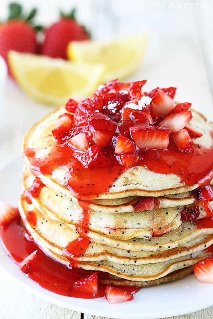 strawberry lemon poppyseed pancakes with strawberry sauce and strawberries on top on a plate with strawberries and slices of lemons faded in the background.