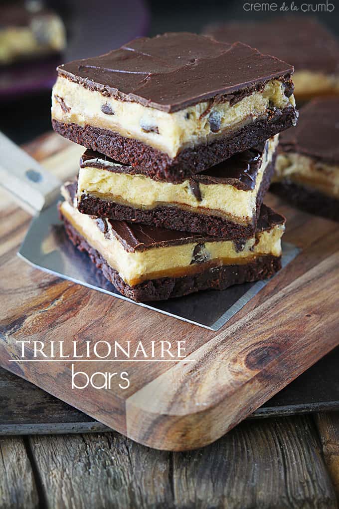 stacked trillionaire bars on a serving spatuala on a cutting board with the title of the recipe written on the bottom left corner of the image.