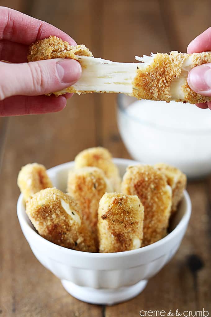 a hand pulling a part a oven baked mozzarella stick above other mozzarella sticks in a bowl with ranch in a bowl with ranch in a bowl faded in the background.