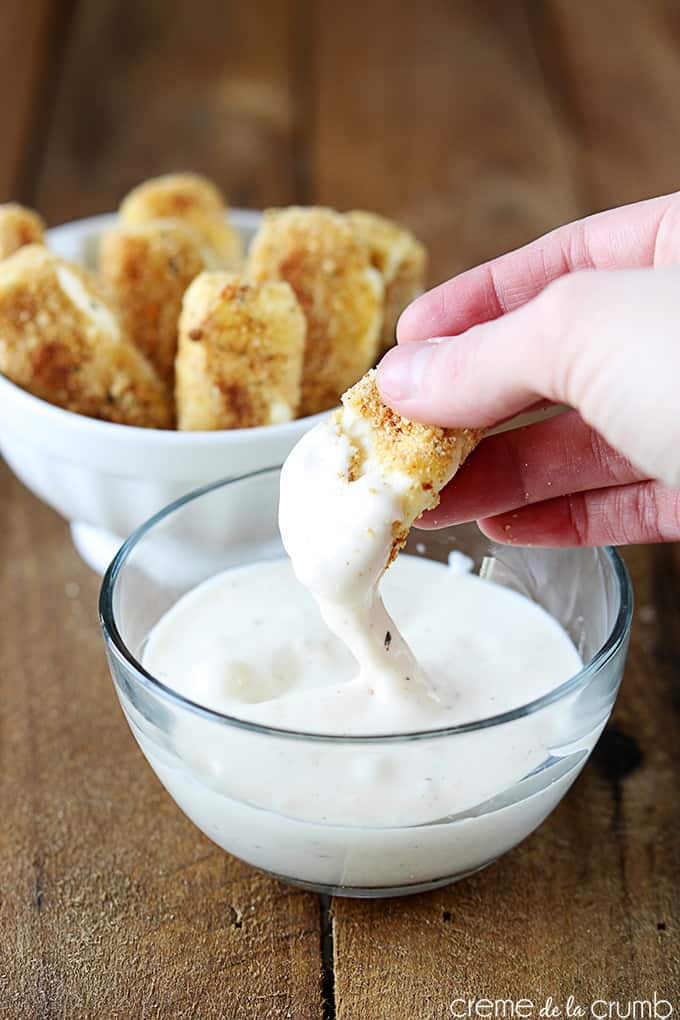 a hand dipping a oven baked mozzarella stick in ranch with more mozzarella sticks in a bowl in the background.