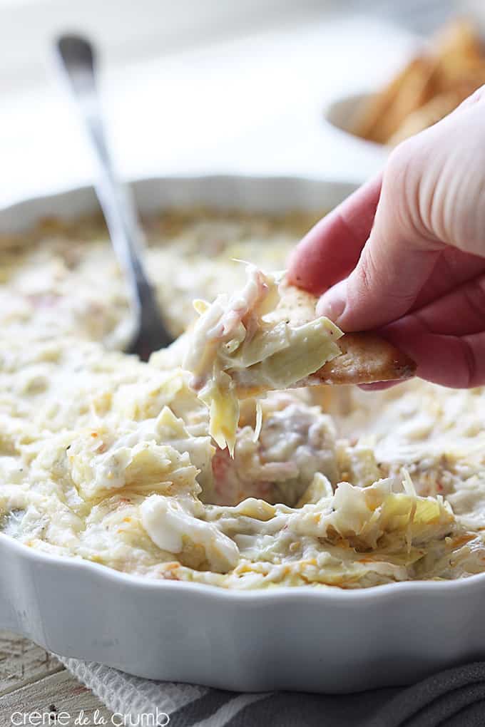 a hand taking a chip with hot crab and cheesy artichoke dip on it from a serving bowl.