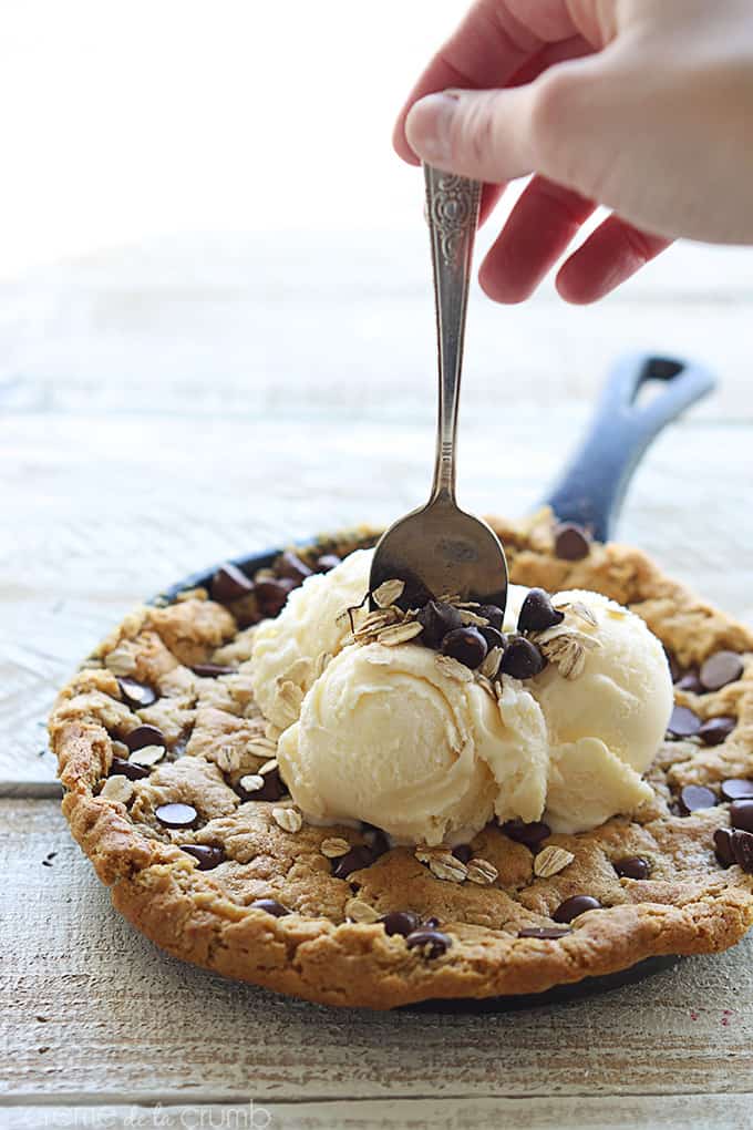 peanut butter oatmeal chocolate chip skillet cookie in a skillet topped with vanilla ice cream, chocolate chips, and oats with a hand holding a spoon at the top of the ice cream.