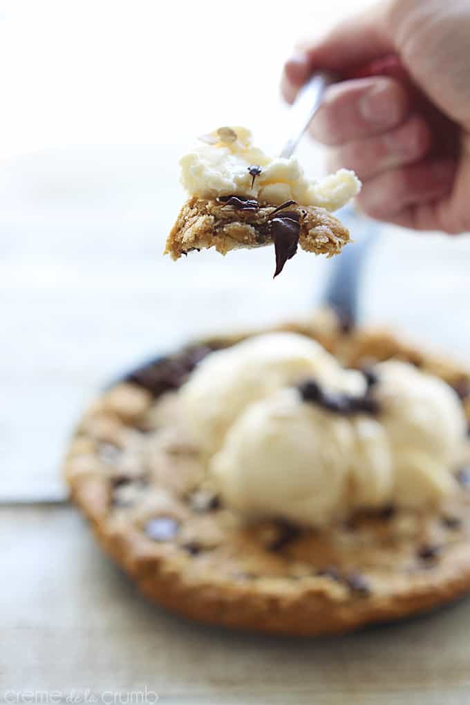 a hand holding a spoon with a bite of vanilla ice cream and peanut butter oatmeal chocolate chip skillet cookie with peanut butter oatmeal chocolate chip skillet cookie in a skillet topped with vanilla ice cream, chocolate chips, and oats faded in the background.