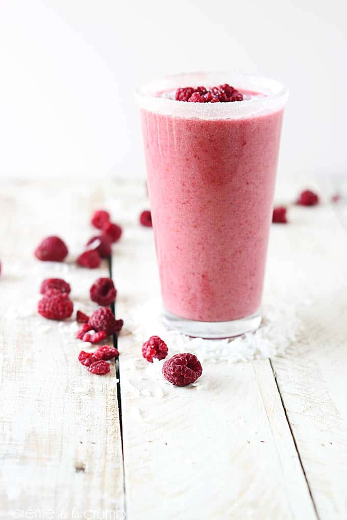a raspberry coconut smoothie in a tall glass on a wooden table with raspberries and coconut shavings.