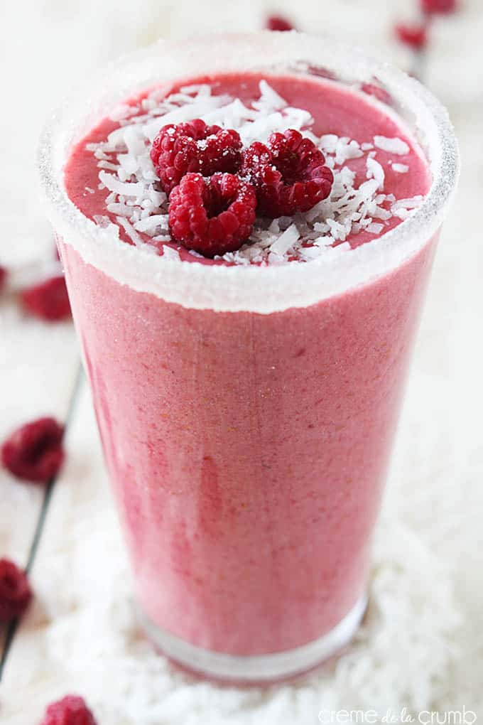 close up of a raspberry coconut smoothie in a tall glass on a wooden table with raspberries and coconut shavings.