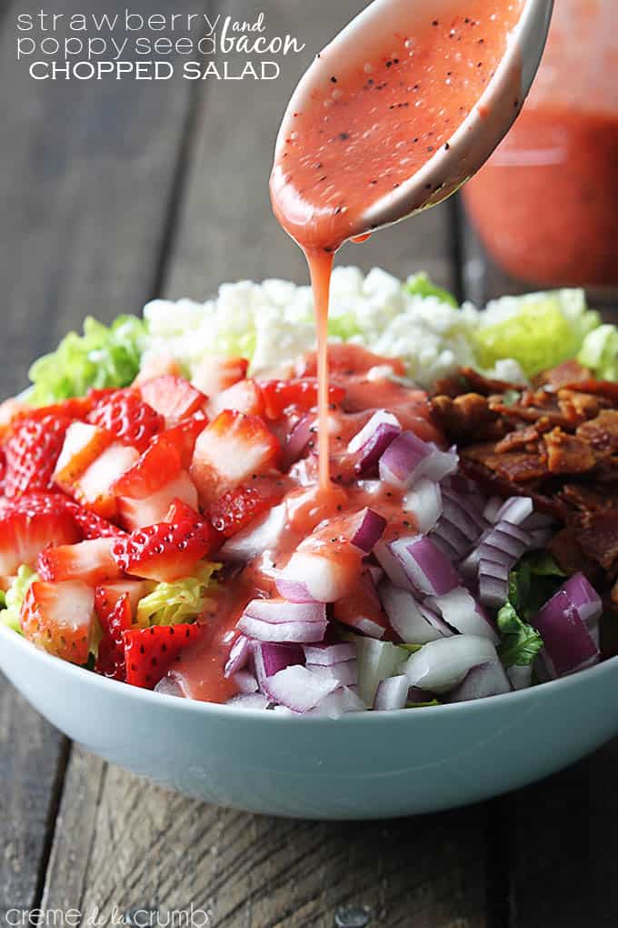 strawberry poppyseed & bacon chopped salad in a bowl with strawberry poppyseed dressing being poured on top with a spoon with the title of the recipe written on the top left corner of the image.
