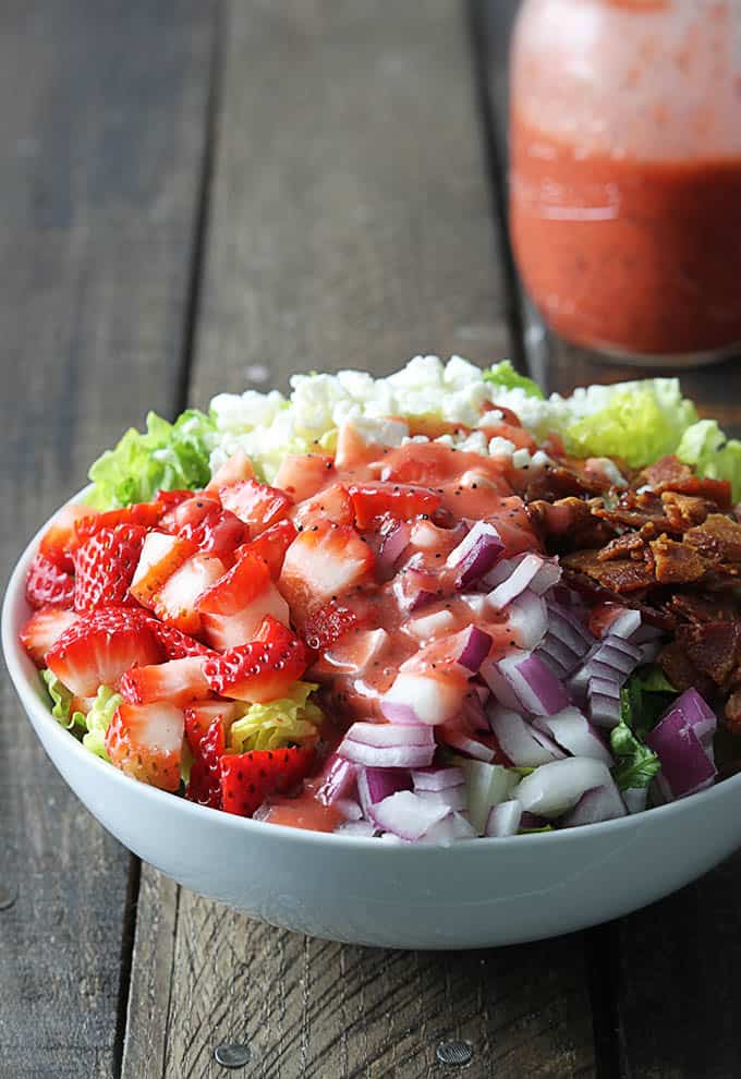 strawberry poppyseed & bacon chopped salad in a bowl with a mason jar of strawberry poppyseed dressing faded in the background.