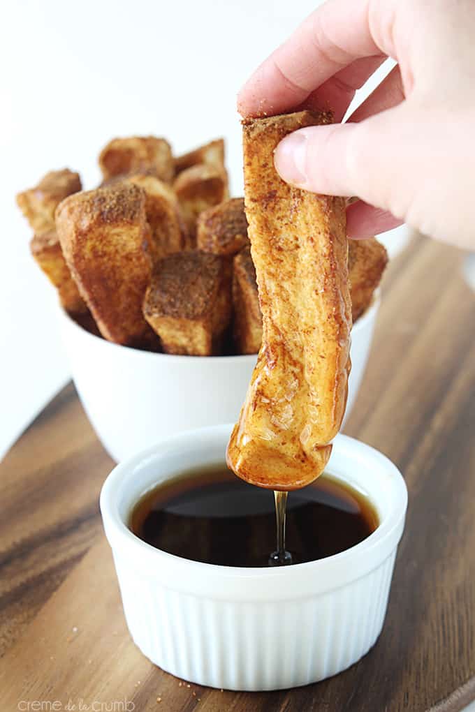 a hand pulling a cinnamon French toast stick out of syrup with more French toast sticks in the background.
