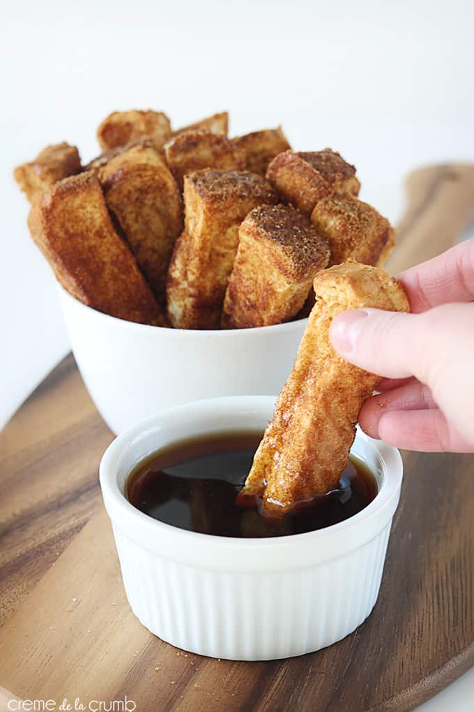 a hand dipping a cinnamon French toast stick in syrup with more French toast sticks in a bowl in the background.