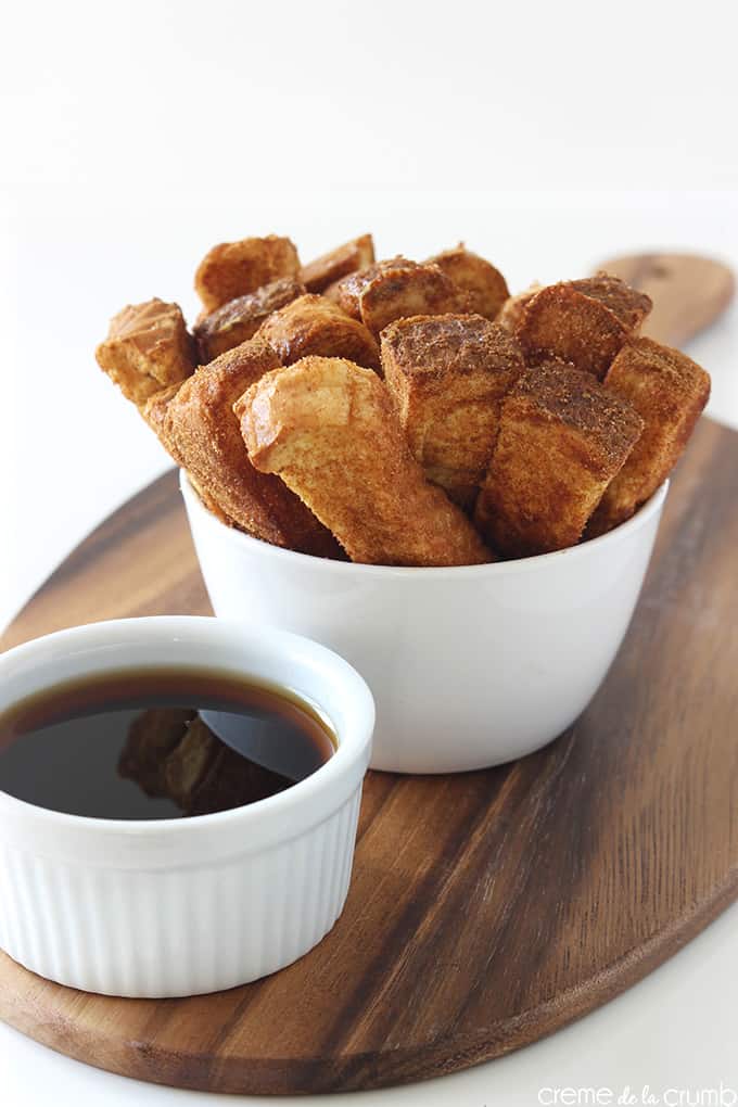 syrup in a dipping bowl with cinnamon French toast sticks in a bowl in the background on a wooden cutting board.