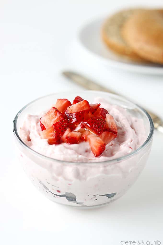 strawberry cream cheese shmear in bowl topped with chopped strawberries with a knife and a bagel on a plate in the background.