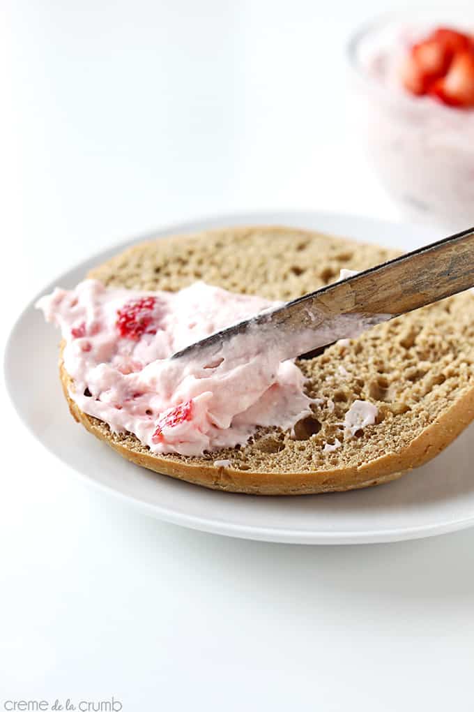 a knife spreading strawberry cream cheese shmear on a bagel on a plate with more shmear in a bowl faded in the background.
