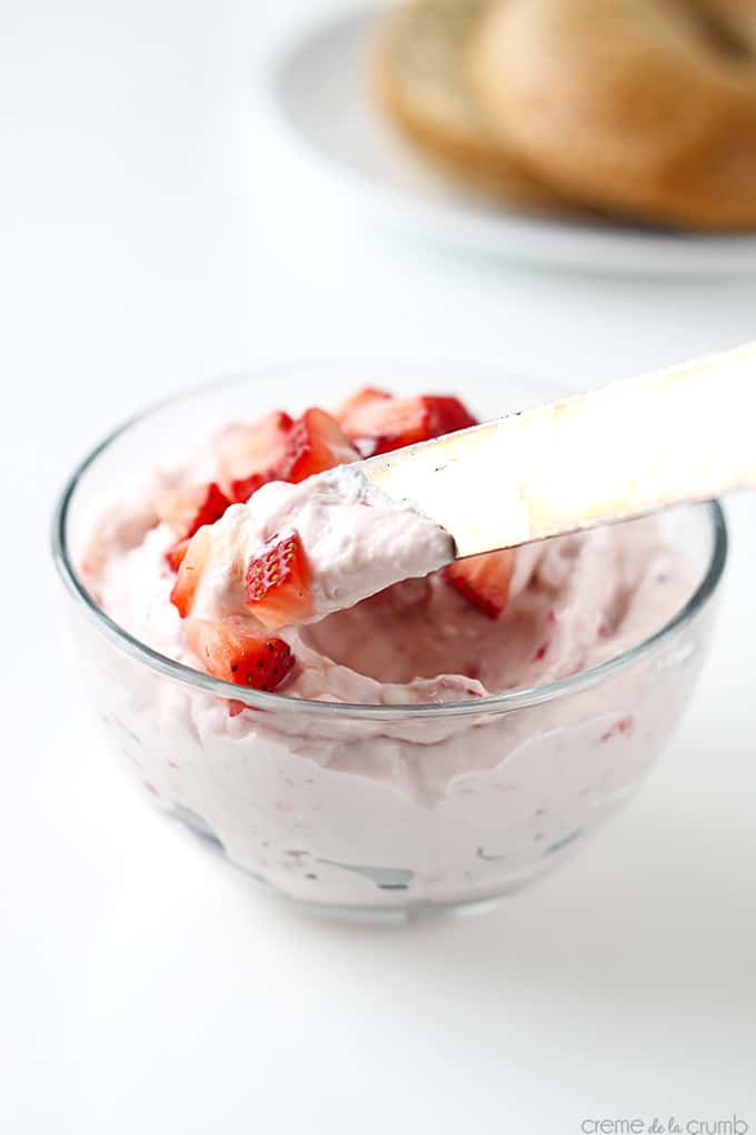 a knife dipping into strawberry cream cheese shmear in a bowl with chopped strawberries with a bagel on a aptly faded in the background.